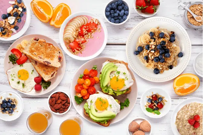 A variety of Easy breakfast foods including a fried egg with toast, a bowl of yogurt with blueberries and granola, a smoothie bowl with granola and fruit, and a glass of orange juice.