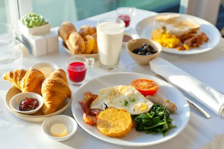 A breakfast table set with various dishes including croissants, fried eggs, hash browns, sautéed greens, grilled tomato, sausage, bacon, a glass of milk, and small bowls of jam and butter.