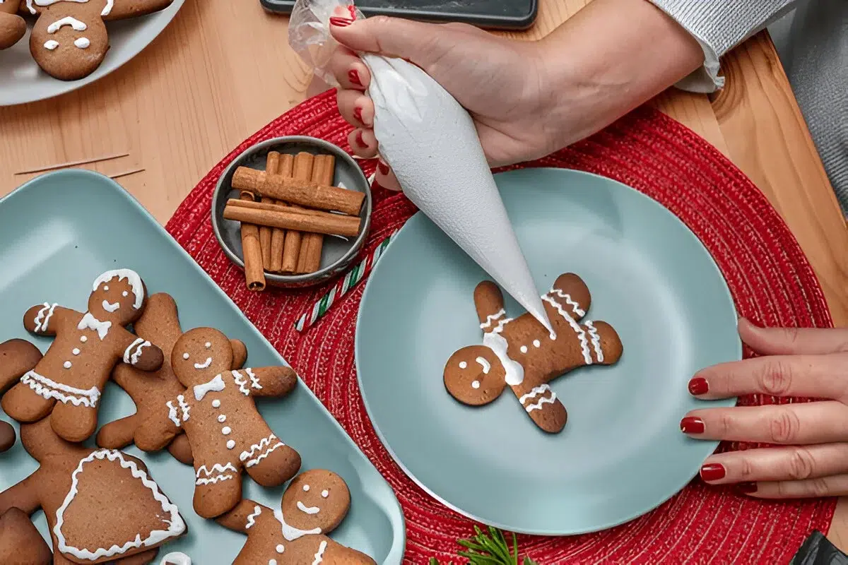 A person decorating a gingerbread cookie with white icing on a blue plate, surrounded by other decorated gingerbread cookies and cinnamon sticks. The scene is part of Christmas Holiday Dessert Ideas.