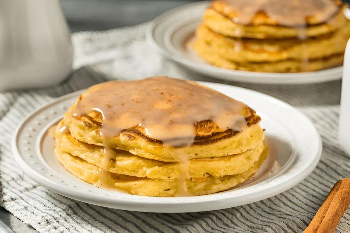 A stack of Eggnog Pancakes topped with a creamy glaze on a white plate. This scene is part of Eggnog Pancakes Christmas Holiday Dessert Ideas.