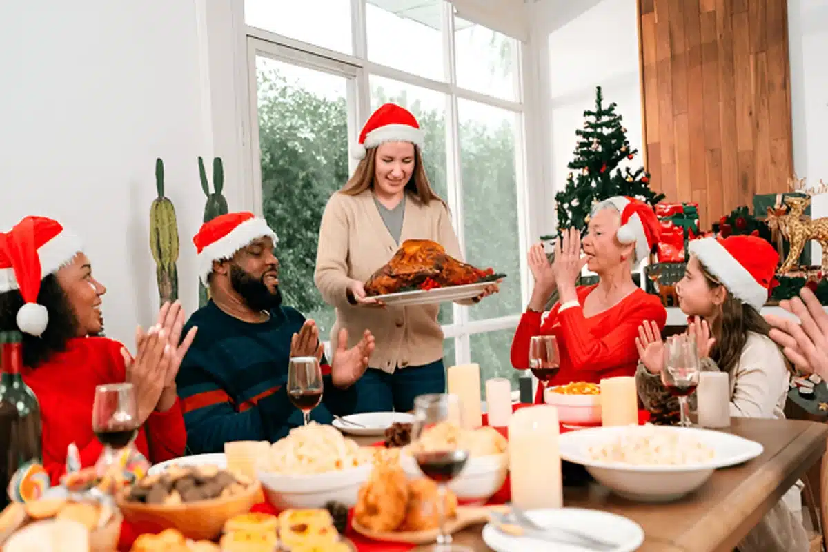 Christmas Dinner Ideas Celebration: People in Santa hats enjoy a festive feast. One presents a roasted turkey as others clap, with a decorated tree in the background.