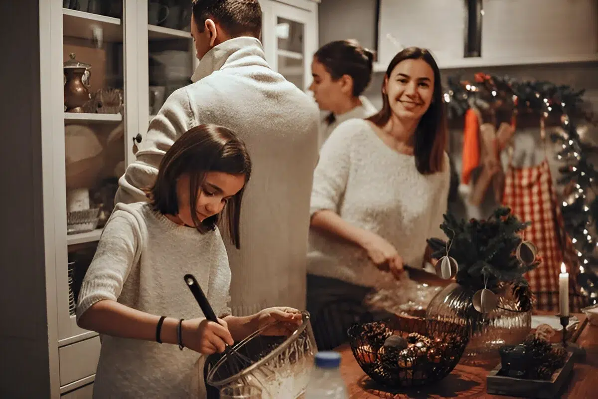 A group preparing food in a festive kitchen, with one whisking ingredients and another handling plastic wrap, surrounded by Timeless Christmas Dishes and holiday decorations.