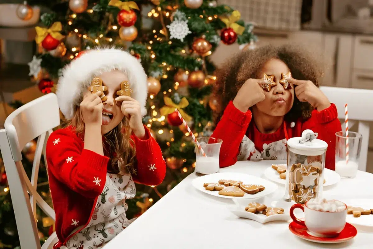 Christmas Desserts :Two children in red sweaters and festive aprons sit at a table with Christmas cookies and milk, with a decorated Christmas tree in the background