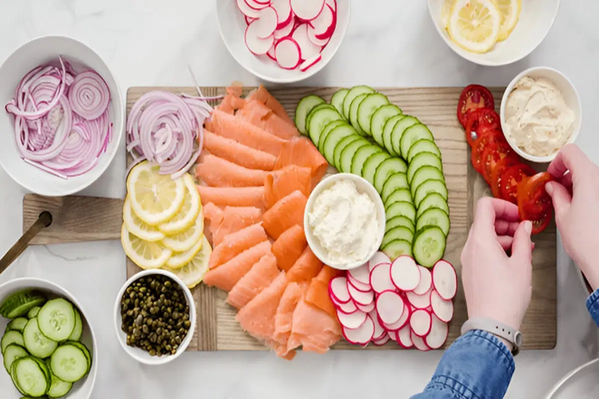 A wooden board with an assortment of sliced vegetables, smoked salmon, and various toppings, surrounded by bowls containing additional ingredients.