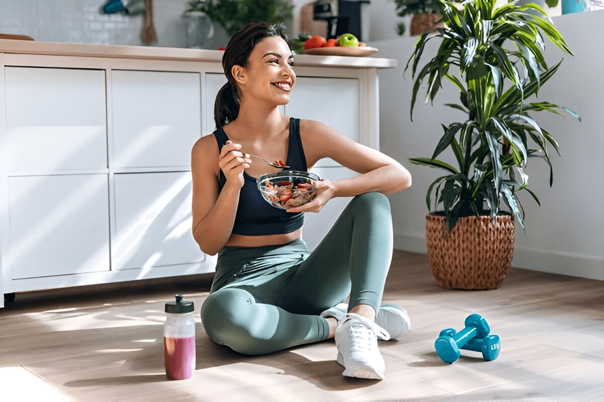 A person in athletic wear holding a bowl of a healthy breakfast, with a water bottle and dumbbells on the floor, and fruits on a kitchen counter.