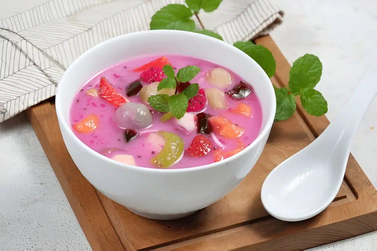 A bowl of vibrant pink "healthiest desserts" soup with various colorful fruits and jelly pieces, garnished with a sprig of mint, placed on a wooden tray with a white spoon beside it